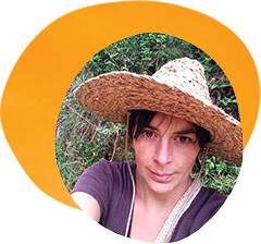 Andrea Echeverri, a white woman with brown hair, poses in front of a patch of woods. She wears a straw hat and a gray shirt.