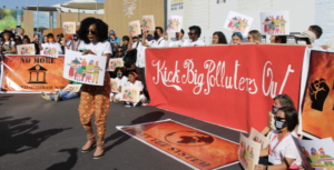 People gather outside with a red banner that read "Kick Big Polluters out!" A Black woman with curly hair stands in front of the crowd, holding a sign with the same demand, and leads everyone in song.