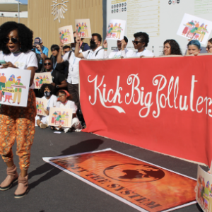 People gather outside with a red banner that read "Kick Big Polluters out!" A Black woman with curly hair stands in front of the crowd, holding a sign with the same demand, and leads everyone in song.