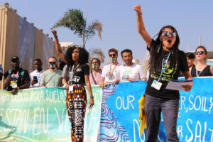 Two women, stand in front of a group of activists and lead a protest chant. They raise their fists in the air, channeling their energy and passion for climate justice.