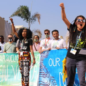 Two women, stand in front of a group of activists and lead a protest chant. They raise their fists in the air, channeling their energy and passion for climate justice.