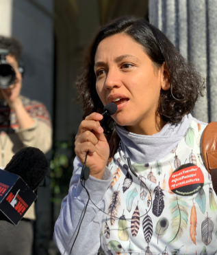 Nathalie Refingo Alverez speaks with passion a microphone at a press conference. She has short brown hair, light-brown skin, and wears a "Make Polluters Pay" pin on her sweatshirt.