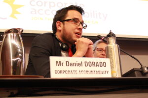 Daniel Dorado speaks into a microphone at a panel discussion. He has dark hair, pale skin, and wears classes, and sits in front of a presentation screen with the Corporate Accountability logo.