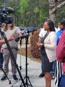 A Black woman, dressed in a beige sweater and black, striped dress is interviewed by the media. She is outside, facing a crew holding audio and visual equipment. Her community stands behind her.