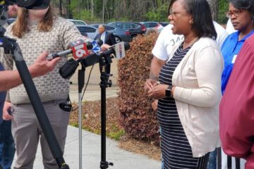 A Black woman, dressed in a beige sweater and black, striped dress is interviewed by the media. She is outside, facing a crew holding audio and visual equipment. Her community stands behind her.