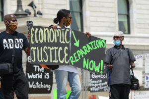 A Black woman, wearing a brightly-colored fabric face mask, stands outside of the Minnesota capital building holding protest signs that read "Economic justice" and "reparations now!" She is surrounded by Black men holding video equipment and participating in the protest.