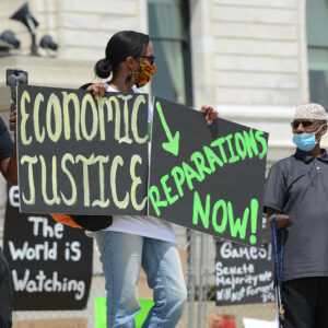 A Black woman, wearing a brightly-colored fabric face mask, stands outside of the Minnesota capital building holding protest signs that read "Economic justice" and "reparations now!" She is surrounded by Black men holding video equipment and participating in the protest.