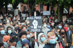 A group of people gather in protest. Many wear protective face asks. A person in the middle holds a sig that reads "All power to the people' with an image of a fist.
