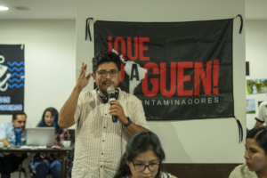 Martin Vilela, climate campaign organizers, speaks into a microphone to a crowd of organizers. Martin has dark hair, light brown skin, and glasses. He wears a pin-striped shirt and headphones rest on his neck. Behind him, a black and red banner reads: Que Paguen! - the campaign's slogan.