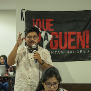Martin Vilela, climate campaign organizers, speaks into a microphone to a crowd of organizers. Martin has dark hair, light brown skin, and glasses. He wears a pin-striped shirt and headphones rest on his neck. Behind him, a black and red banner reads: Que Paguen! - the campaign's slogan.