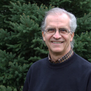 Portrait of Dan Crawford. Dan has white skin and hair and glasses. He wears a navy blue sweater, stands in front of pine trees, and smiles.