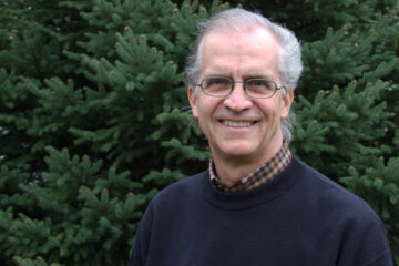 Portrait of Dan Crawford. Dan has white skin and hair and glasses. He wears a navy blue sweater, stands in front of pine trees, and smiles.
