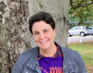 Portrait photo of Anna Mudd. Anna has short brown hair and she's wearing a gray jacket over a purple shirt. She's standing in front of a tree.