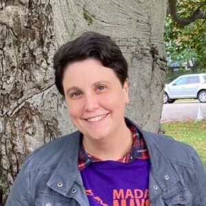 Portrait photo of Anna Mudd. Anna has short brown hair and she's wearing a gray jacket over a purple shirt. She's standing in front of a tree.