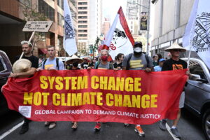 Crowds gather and march down the streets of New York City demanding climate justice. Five people of different ages and backgrounds carry a banner that reads "System change not climate change."