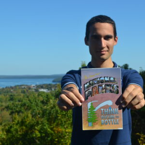 : Ari Rubenstein, a young white organizer with Corporate Accountability, holds a sign that says “National Parks Think Outside the Bottle.” He is high above the scenic background behind him--blue sky, ocean, and trees.