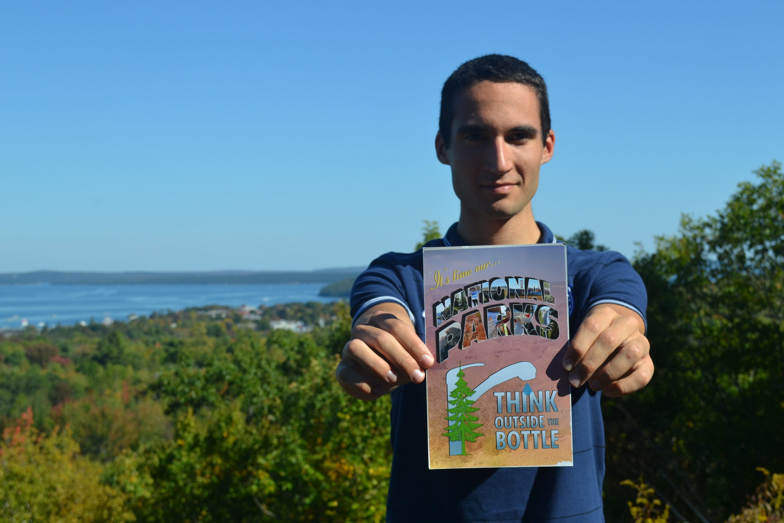 : Ari Rubenstein, a young white organizer with Corporate Accountability, holds a sign that says “National Parks Think Outside the Bottle.” He is high above the scenic background behind him--blue sky, ocean, and trees.