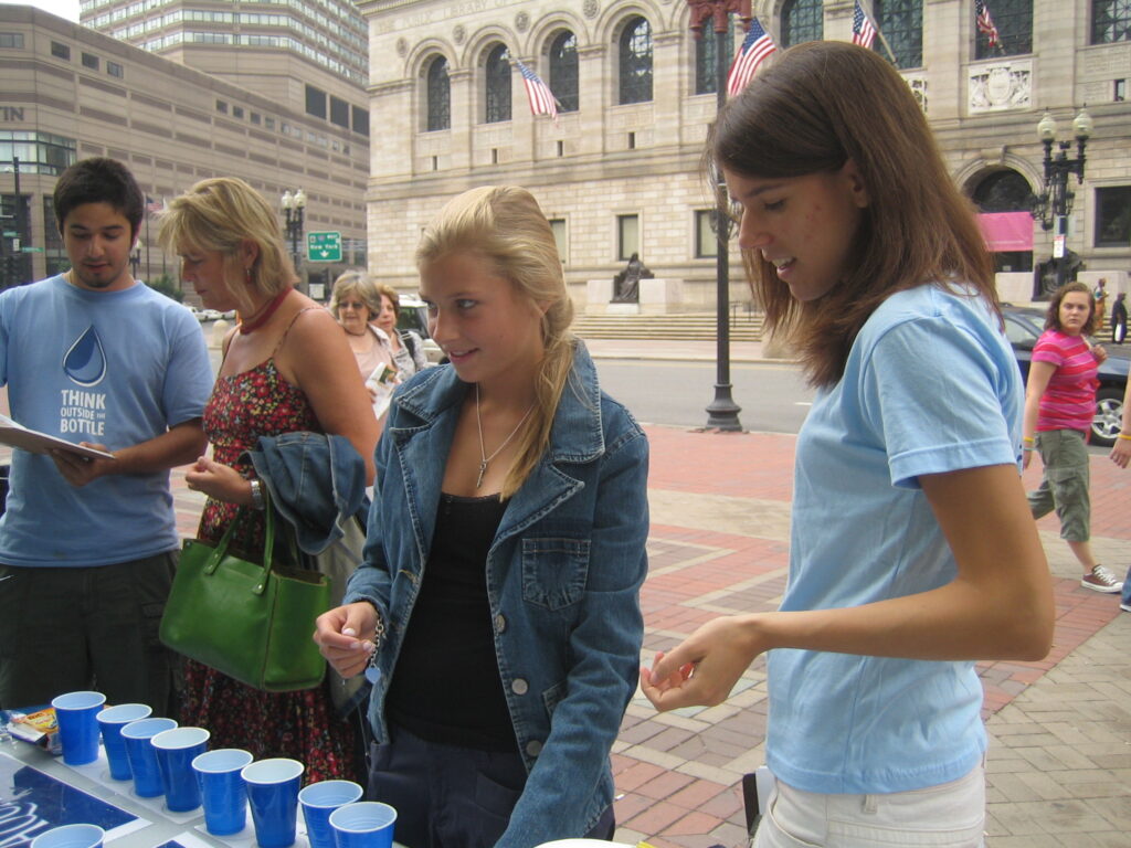 A photo of two people standing in front of a table filled with blue cups, about to take the Tap Water Challenge. An organizer on the left holds a clipboard and talks to a passer-by.