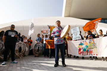 A group of people gather to protest outside of the U.N. climate talks. An Indigenous leader stands before the crowd and with a handheld drum and leads the group in song.