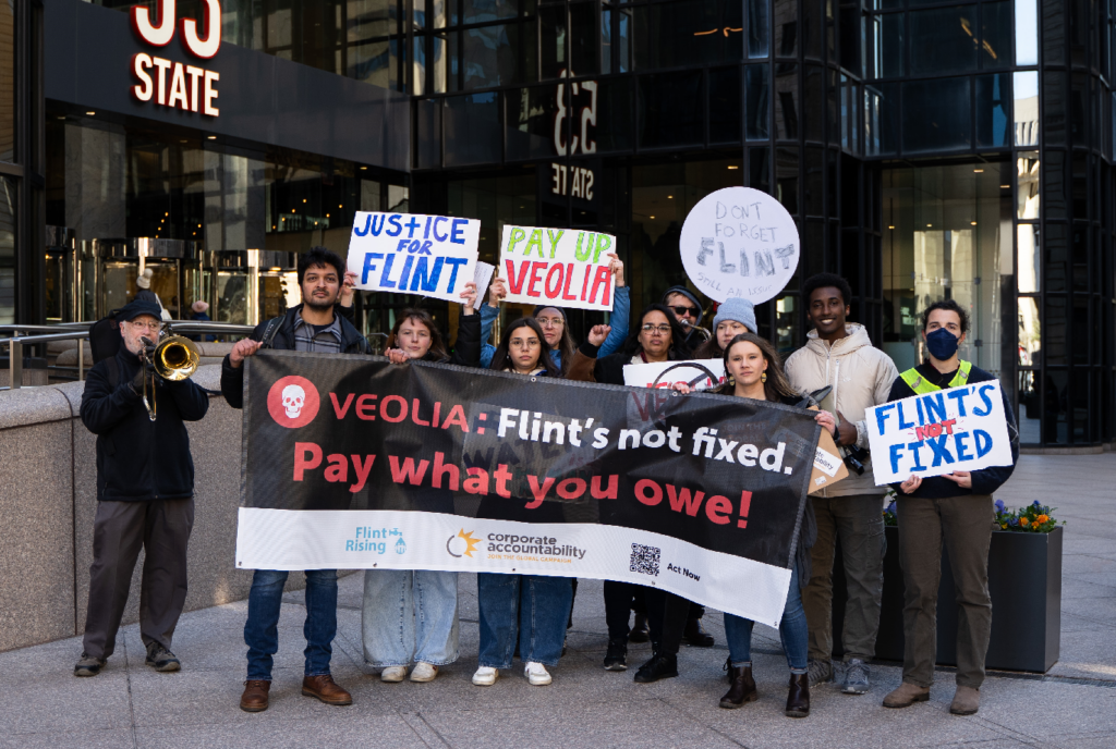 A group of people stand in front of an office building in downtown Boston, holding signs and a banner that reads: "Veolia, Flint's not fixed. Pay what you owe!"