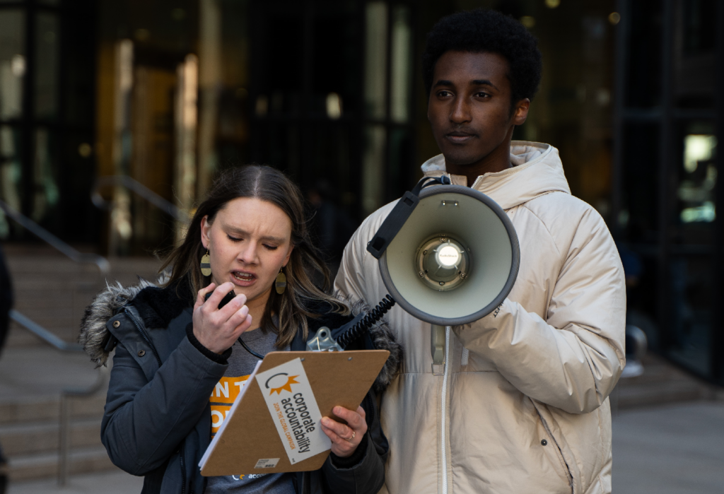 A white woman and a Black man stand together at a rally. The woman holds a clipboard and speaks into a microphone. 