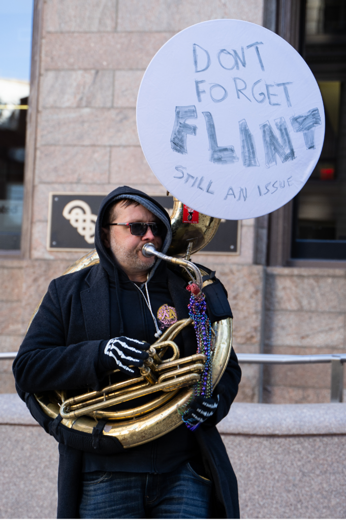A man plays a brass instrument on the streets of Boston. "Don't forget Flint" is written on the bell.