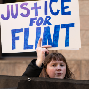 A white woman with light brown hair holds up a sign that reads "Justice for Flint"