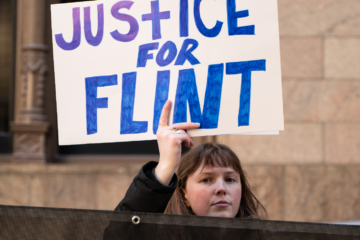 A white woman with light brown hair holds up a sign that reads "Justice for Flint"