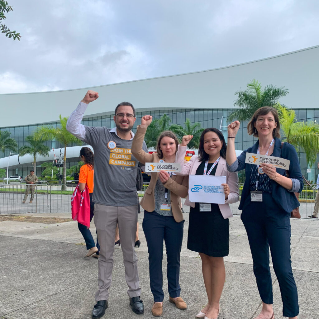 Four Corporate Accountability organizers stand outside of a large building venue that is surrounded by palm trees. They hold signs and wear shirts that read "Corporate Accountability" and "Join the Global Campaign" and raise their fists in the air.