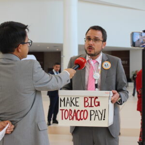 Organizer Daniel Dorado speaks to a media person outside of the negotiating halls. He is holding a large white box that reads "Make Big Tobacco Pay!"