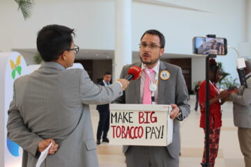 Organizer Daniel Dorado speaks to a media person outside of the negotiating halls. He is holding a large white box that reads "Make Big Tobacco Pay!"
