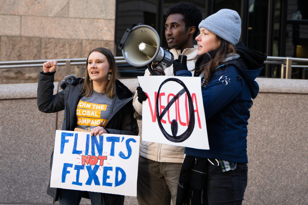 Three organizers--two white women and a Black man--stand in the cold outside a city building. One has her fist in the air. Another speaks into a megaphone. Their signs link Veolia to its role in Flint.