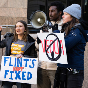 Three organizers stand together outside of a city building holding signs that protest Veolia for its role in Flint and speaking into a mega phone.