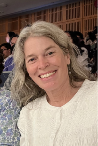 Joanna, a white woman with long grayish blondish hair smiles at the camera. She's wearing a white shirt.