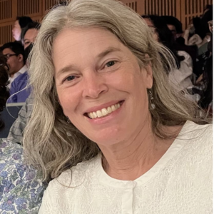 Joanna, a white woman with long grayish blondish hair smiles at the camera. She's wearing a white shirt.