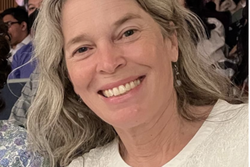 Joanna, a white woman with long grayish blondish hair smiles at the camera. She's wearing a white shirt.