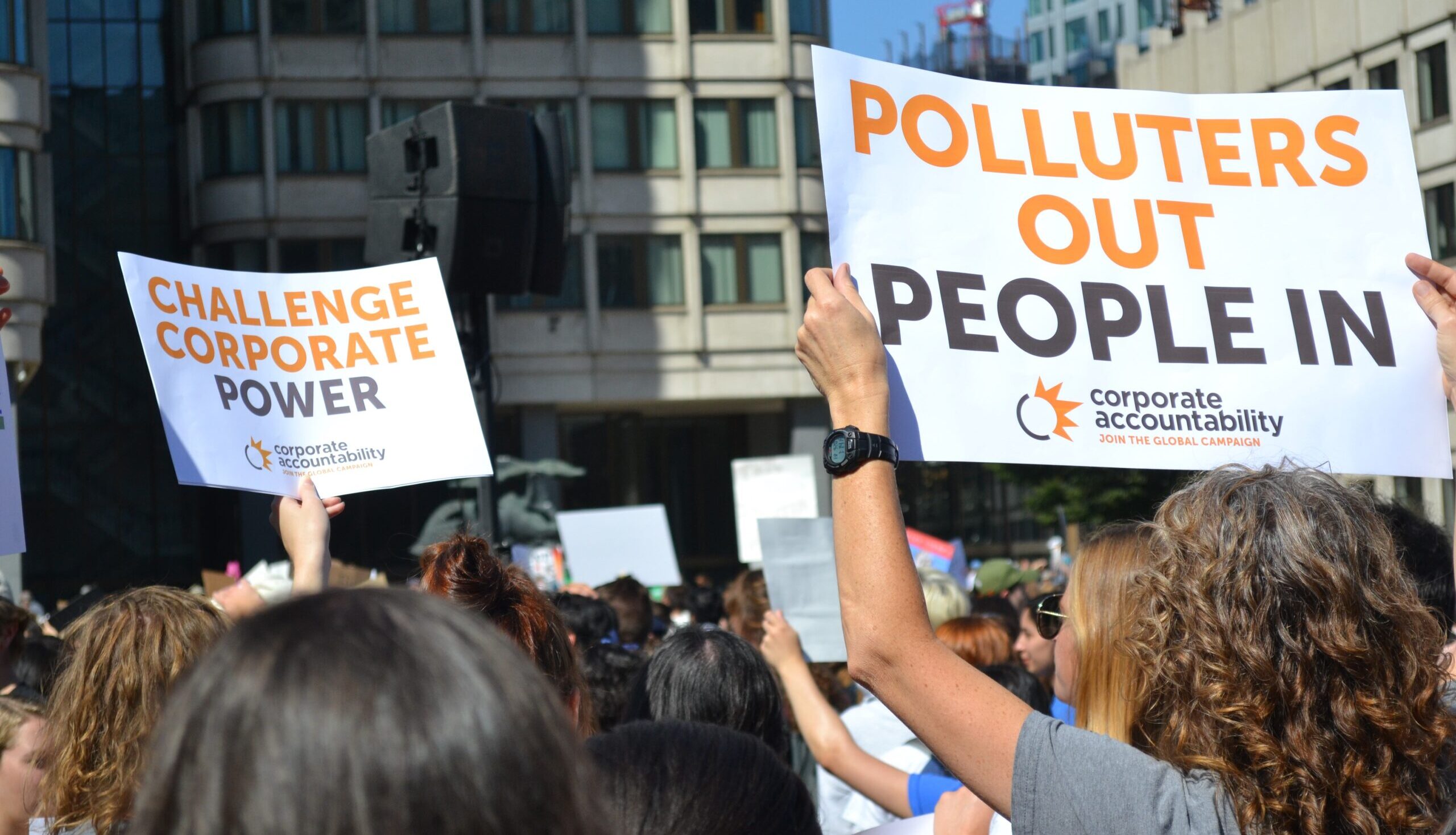 Hundreds of people gather at a protest and hold signs that read "Challenge Corporate Power" and "Polluters Out People In!"