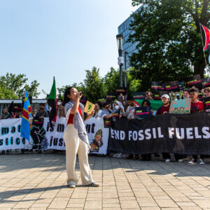 A woman speaks into a megaphone at a protest. People in the background hold signs that read end Fossil Fuels Now.