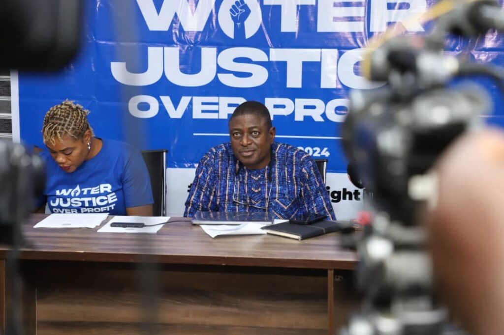 A close up shot of Akinbode Olufemi, sitting at the panelist's table. Bode is a Black man. He is wearing a blue shirt and is looking straight ahead, toward the crowd.