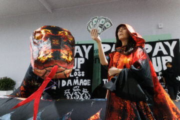 A woman in a orange and black cape holds up a fan of dollar bills, next to a giant blow up snake. Behind her, people hold signs that read "pay up!"