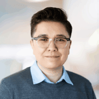 Head shot of Ari Belathar. They have dark brown, short hair, and they are wearing glasses and a gray sweater over a blue collared shirt.