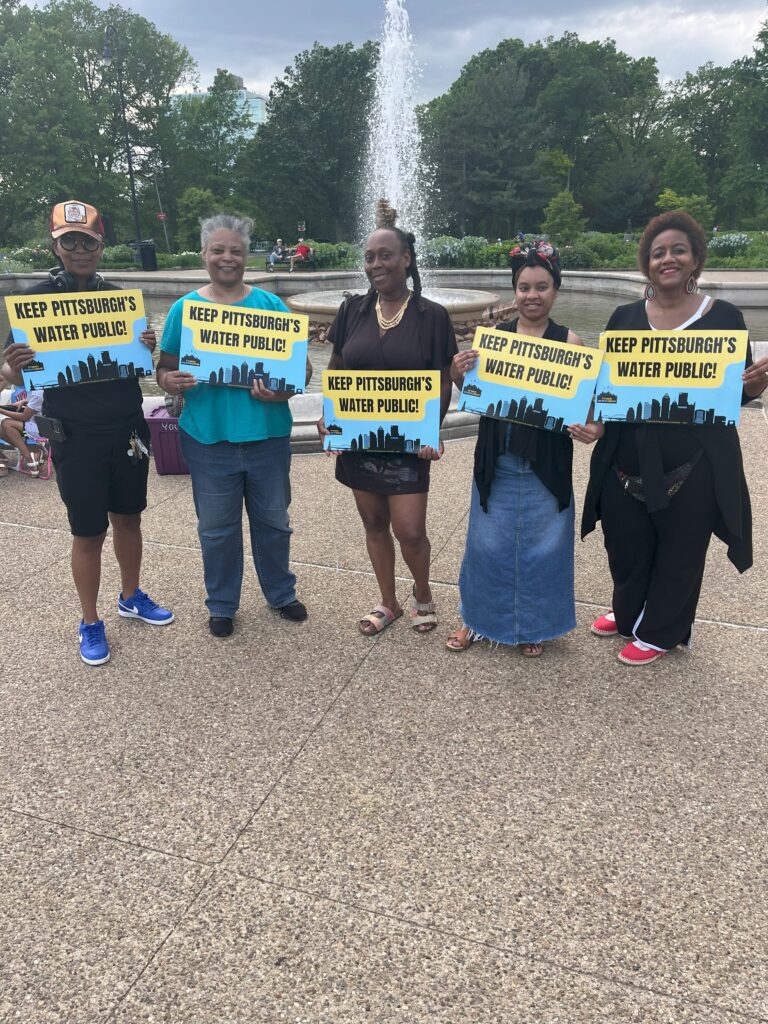 Five Black women stand together in front of a water foundation holding signs that read "Keep Pittsburgh's water public."