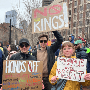 Three friends at mobilization challenging rise of fascism in the U.S. holding signs that read "no kings!" and "people over profit"