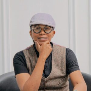 Portrait of Gabby Gray. She wears a gray cap and a vest, rests her chin in her hand, and smiles at the camera.