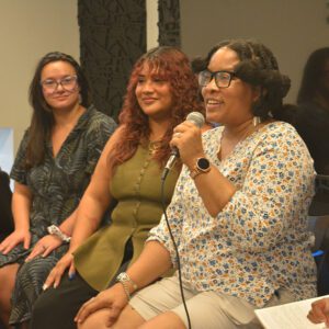 Three people engaging in a panel discussion, with the person closest to the camera talking into the microphone.