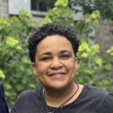 Portrait of Onah Ruth Assai, smiling at the camera, waring a black shirt and leather necklace.