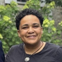 Portrait of Onah Ruth Assai, smiling at the camera, waring a black shirt and leather necklace.