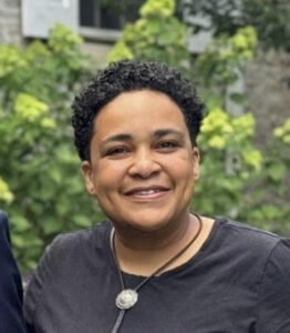 Portrait of Onah Ruth Assai, smiling at the camera, waring a black shirt and leather necklace.